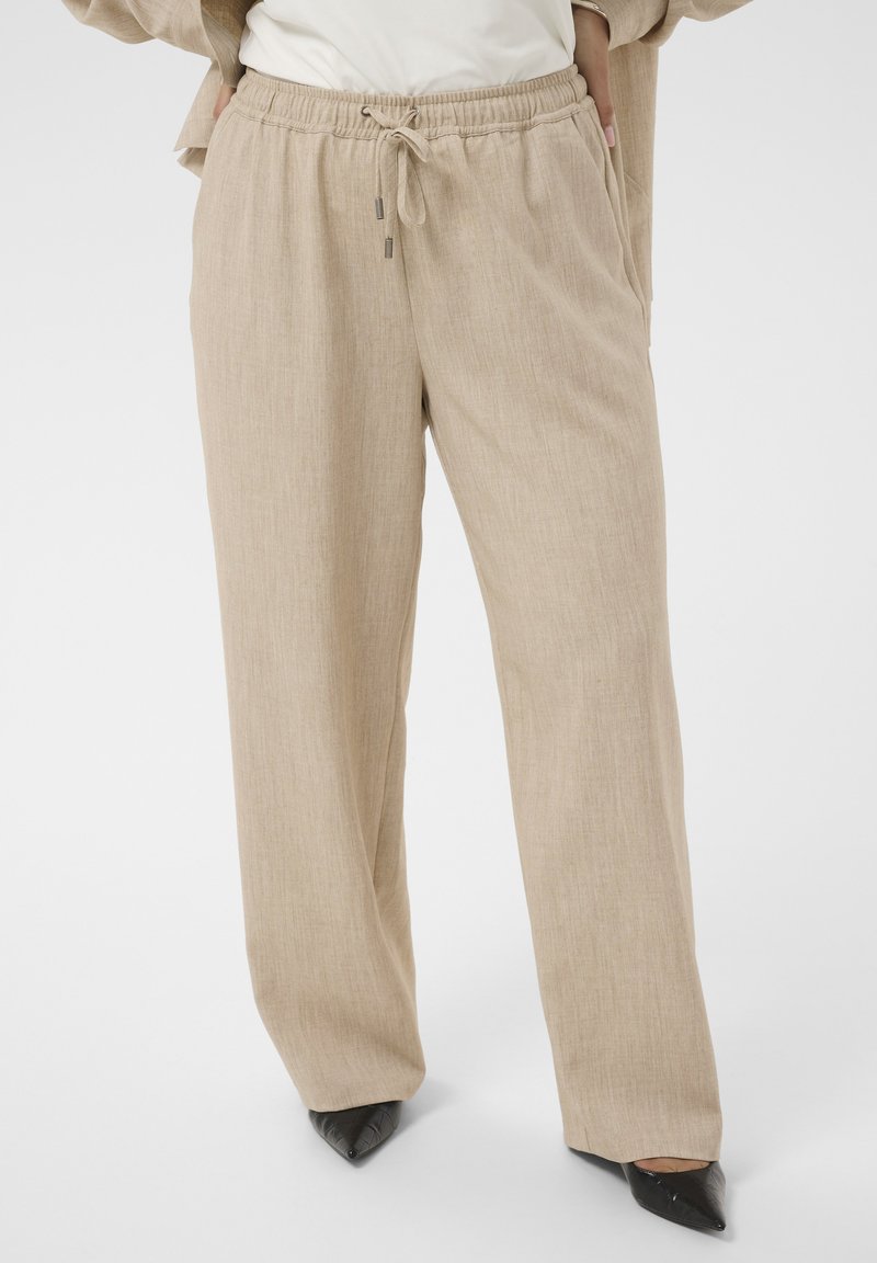 Pantalon beige à jambes larges avec taille élastique et cordon, porté avec des chaussures noires pointues et un haut blanc partiellement visible.