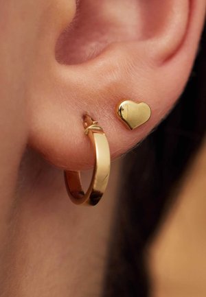 Goldohrringe und kleine Herzstecker, beide glänzend, vor einem Nahaufnahme eines menschlichen Ohrs mit heller Hautfarbe und feiner Haarstruktur.