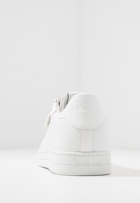 Zapatilla de cuero blanco con acabado texturizado, forma redondeada, logo en relieve en el talón y suela de goma lisa. Diseño minimalista sin patrones visibles.