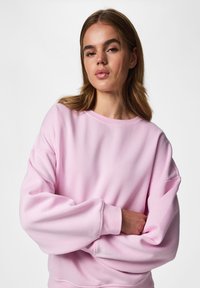 Jeune femme aux cheveux châtain clair portant un sweat-shirt ample rose clair, debout les bras croisés devant un fond uni.