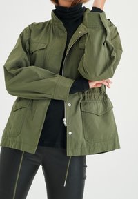 Olive green jacket with a zip front, elastic waist, and two front pockets. Worn over a black turtleneck, with a smooth, lightweight fabric.