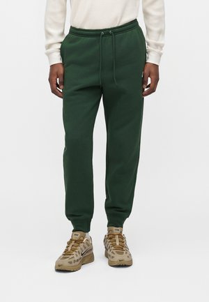 Persoon draagt donkergroene joggingbroek met trekkoord in de taille, beige en bruine Nike-sneakers, en een crèmekleurige longsleeve met structuur tegen een effen achtergrond.