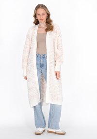 Cardigan lung, cu tricotaj deschis, în crem, cu accente piersică, având manșete și tiv cu elastic. Asortat cu un top bej și blugi evazați de culoare albastră deschis.