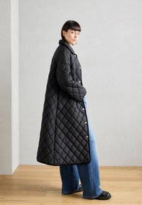 Svart quiltet jakke med diamantmønster, med høy krage og trykknapper. Kombinert med vide blå jeans og svarte sko.