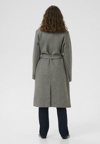 Donna con capelli ricci che indossa un cappotto di lana grigio con cintura, pantaloni scuri e scarpe nere, in piedi con le spalle rivolte verso l'osservatore su uno sfondo bianco.