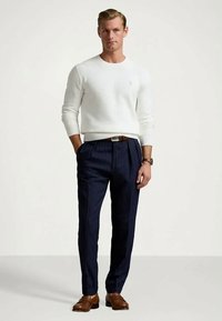 Man i vit tröja, marinblå randiga byxor, brun bälte och skor står med en hand i fickan mot en enfärgad bakgrund.