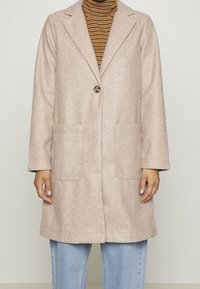 Beige kabát na jedno knoflíček s klopami a velkými předními kapsami, oblečený přes hnědo-černý pruhovaný rolák a světle modré džíny.