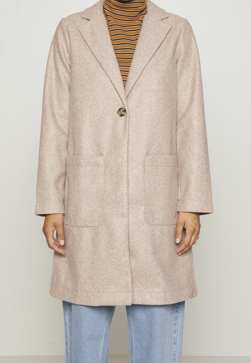 Beige kabát na jedno knoflíček s klopami a velkými předními kapsami, oblečený přes hnědo-černý pruhovaný rolák a světle modré džíny.