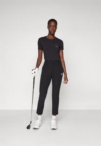 Zwart golfoutfit met een aansluitend t-shirt met een hartgrafiek en een op maat gemaakte broek. Draagt witte schoenen en houdt een golfclub vast.