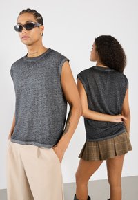Gray, sleeveless t-shirt in a textured fabric worn with beige trousers. A model behind wears a dark, plaid mini skirt. Both have sunglasses.
