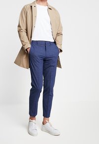 Ljusbeige lång kappa över en vit t-shirt, tillsammans med marinblå skräddarsydda byxor och vita sneakers. Slät tyg med en figursydd design.