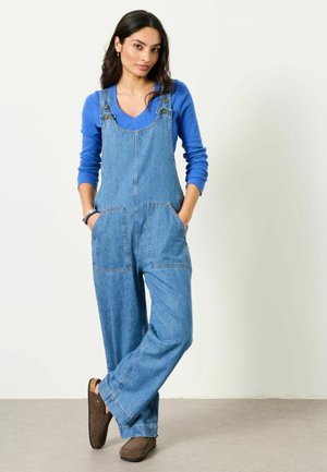 Woman with long dark hair wearing blue long-sleeve shirt, denim overalls, and brown shoes, standing with hands in pockets against plain background.