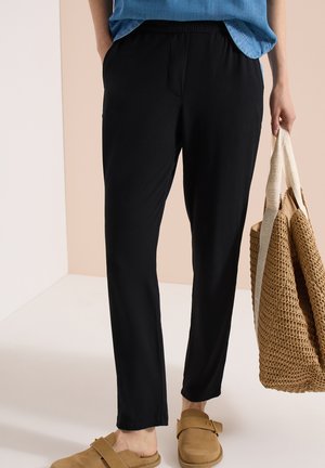 Person trägt schwarze Hosen mit elastischem Bund, hält eine große geflochtene Tasche mit cremefarbenen Trägern und trägt beige Slipper.