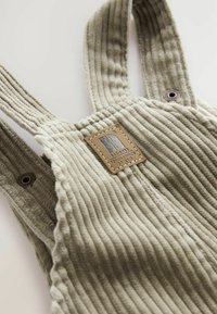 Salopettes en velours côtelé vert olive avec des rayures verticales texturées. Comprend des éléments en métal et un logo en patch en cuir cousu.