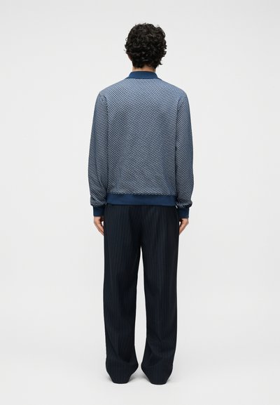 Άνδρας με σγουρά μαλλιά στέκεται με την πλάτη, φοράει μπλε σακάκι με σχέδια και χαλαρά σκούρα παντελόνια με ρίγες, μπροστά σε απλό φόντο.