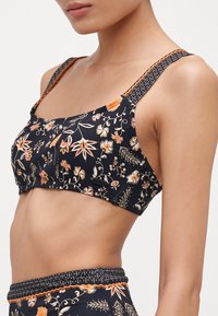Bikini topp med blomsterprint i marineblått med oransje og hvite aksenter. Har brede stropper og teksturert detaljering på stroppene. Laget av stretchstoff.
