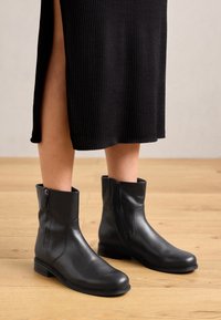 Černé kožené kotníkové boty s hladkou strukturou, bočním zipem a nízkým blokovým podpatkem, doplněné černým žebrovaným oděvem.