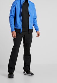 Blauwe softshell-jas met zwarte accenten, met een rits aan de voorkant, gecombineerd met zwarte broek en zwarte sportieve schoenen. Minimalistisch design en een aansluitende pasvorm.