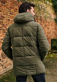 Jachetă căptușită de culoare verde măsline, cu glugă, având un design matlasat, mâneci lungi și o croială ușor prelungită, amplasată pe un perete din cărămidă.