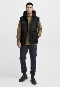 Gilet imbottito nero con cappuccio, dotato di numerose tasche e cerniera. Indossato sopra una maglietta a maniche lunghe marrone e abbinato a pantaloni scuri e stivali.