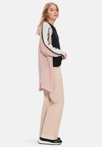 Langer, pinker Strickcardigan über einem schwarzen Shirt, kombiniert mit hellrosa, weit geschnittenen Hosen und schwarzen Sneakers. Kontrastierende gestreifte Pulloverärmel.