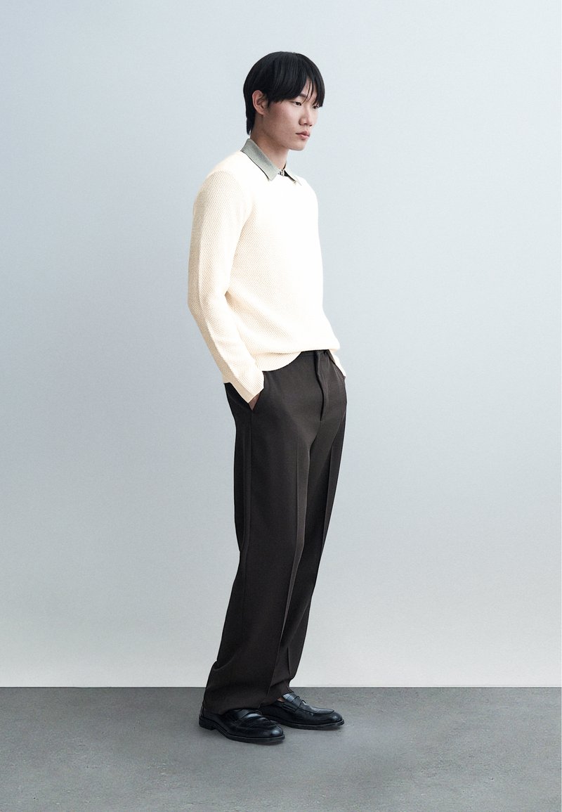 Young man standing with hands in pockets, wearing cream sweater over collared shirt, dark trousers, and black shoes against plain light background.