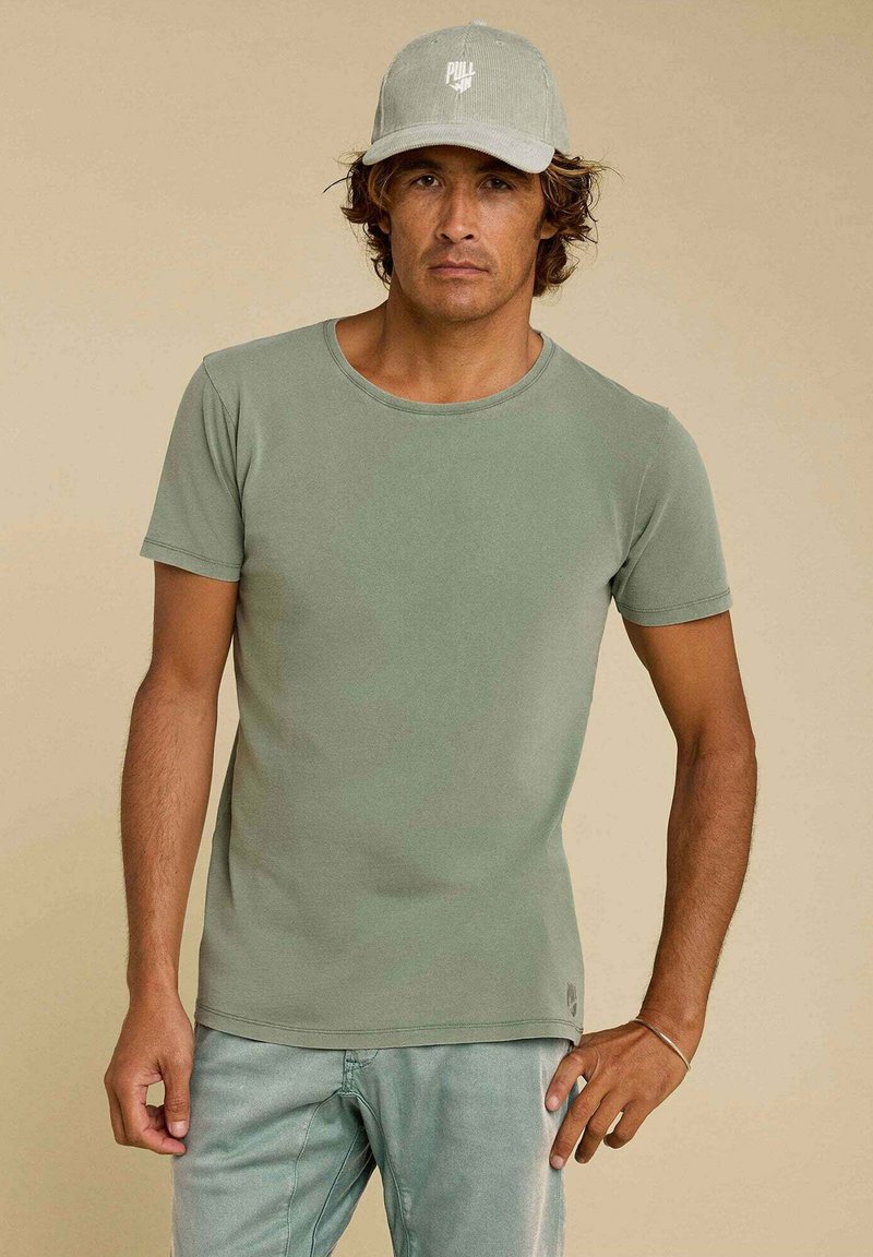 T-shirt à manches courtes et col rond, de couleur vert muted, fabriqué en tissu lisse. Porté avec un pantalon vert clair et une casquette grise avec logo.