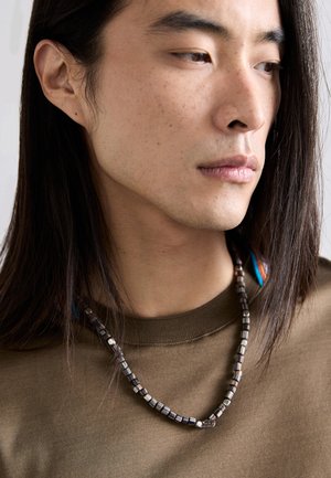 Collana di perle con un mix di perle metalliche scure e argento, indossata su una maglietta marrone con dettaglio a girocollo. Lunghi capelli scuri visibili.