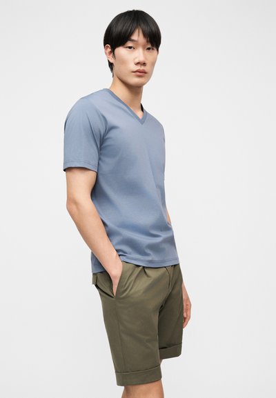 Νέος άνδρας με μαύρα μαλλιά που φοράει μπλε μπλουζάκι με V-λαιμόκοψη και χακί βερμούδα έως το γόνατο, με τα χέρια στις τσέπες.