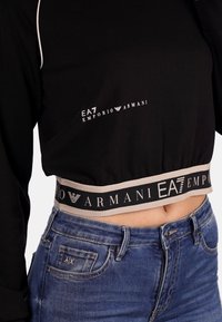Sweatshirt cropped nero con cuciture bianche e logo "EA7 Emporio Armani" sul petto. Presenta un elastico con marchio a contrasto in vita. Jeans blu visibili.