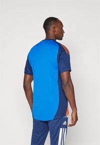 Camisola desportiva azul com painéis laterais escuros e riscas laranja nos ombros, feita de tecido leve e com decote arredondado.