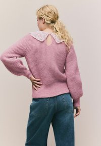 Różowy sweter z dzianiny z koronkowym kołnierzykiem i dekoltem w kształcie łezki z tyłu; zestawiony z niebieskimi dżinsami z tylnymi kieszeniami.