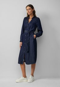 Marineblauwe denim jurk met lange mouwen, knoopsluiting aan de voorkant, tailleband en zijzakken. G gedragen met witte sneakers. Gladde textuur en midi-lengte.