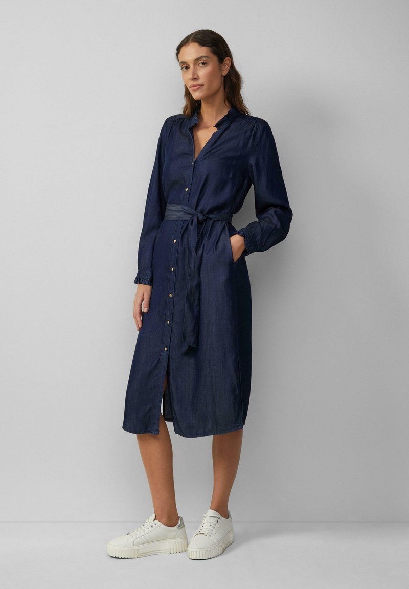 Marineblauwe denim jurk met lange mouwen, knoopsluiting aan de voorkant, tailleband en zijzakken. G gedragen met witte sneakers. Gladde textuur en midi-lengte.