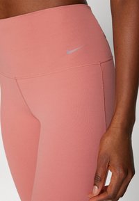 Gros plan sur une personne portant des leggings Nike taille haute rose poussiéreux avec un logo Nike argenté visible près de la hanche.