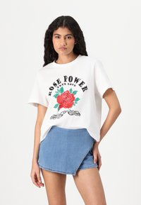 Fehér pamut póló piros rózsa grafikával és fekete felirattal: "ROSE POWER TRUE LOVE"; egy kék farmer mini szoknyával párosítva.
