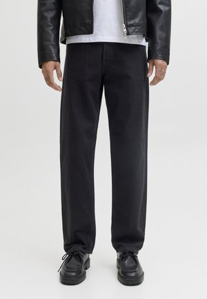 Homme portant un pantalon noir droit, des chaussures en cuir noires, une chemise blanche et une veste en cuir noire sur un fond uni.