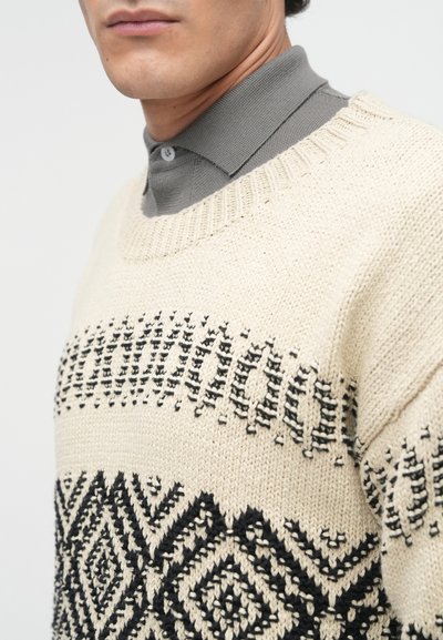 Beige színű kötött pulóver fekete geometrikus mintákkal, kerek nyakkivágással és bordázott díszítéssel. Alatta egy szürke galléros ing található.