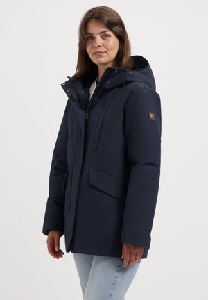 Vrouw in een marineblauwe winterjas met capuchon, met rits en klepzakken, gecombineerd met lichtblauwe jeans, staande tegen een witte achtergrond.