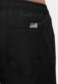 I pantaloni neri presentano una vita elastica, una tasca sul retro con chiusura a bottone e un piccolo etichetta di tessuto all'interno della tasca. Texture liscia.