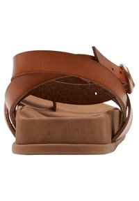 Sandales en cuir marron avec une semelle plate et rectangulaire, ornées de brides croisées et d'une boucle décorative. Texture lisse avec peu de détails.