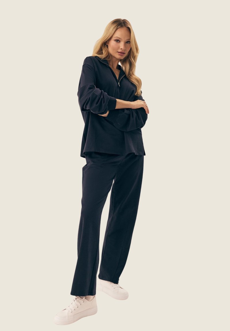 Czarny oversize sweter z kołnierzem na zamek, w połączeniu z dopasowanymi szerokimi spodniami. Białe trampki dopełniają stylizację, podkreślając swobodny wygląd.