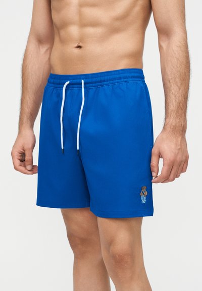 Mężczyzna ubrany w niebieskie kąpielówki z białym sznurkiem i małym haftowanym misiem na lewej nogawce, ukazujący tułów i ramiona.