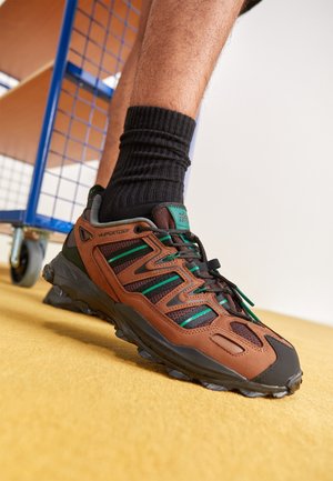 Leg wearing brown and black hiking shoe with green accents and black sock, standing on yellow carpet near blue metal cart wheel.