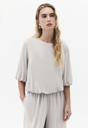 Kvinde med langt blondt bølget hår iført lys grå løs top med puffede halværmer og matchende bukser, kigger let til siden.