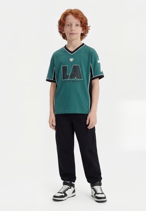 Αγόρι με κόκκινα μαλλιά που φοράει πράσινη μπλούζα με την επιγραφή «LA City of Angels», μαύρο παντελόνι καμπαρντίνα και μαύρα-άσπρα ψηλά αθλητικά παπούτσια, στέκεται πάνω σε λευκό φόντο.