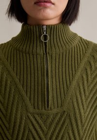 Pull en tricot vert avec un col haut à côtes, présentant une fermeture à demi-zip et des motifs chevrons texturés sur le tissu.