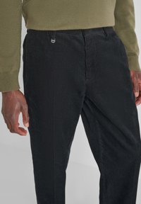 Pantaloni di velluto a coste neri con righe verticali sottili, caratterizzati da una cintura piatta, passanti per la cintura e un dettaglio in metallo. Abbinati a un maglione verde.