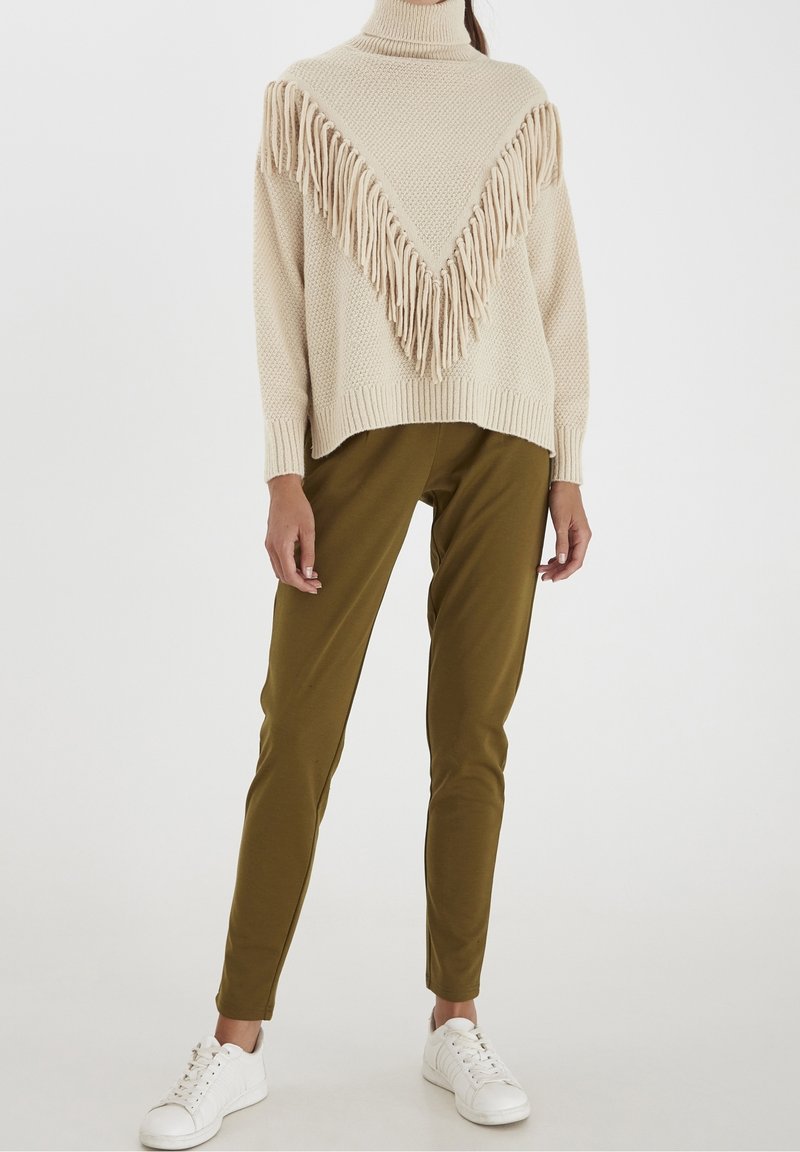Personne portant un pull à col roulé en maille beige à franges, un pantalon vert olive et des baskets blanches, debout devant un fond uni.