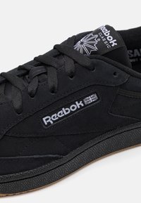 Černé semišové tenisky s texturovaným povrchem a gumovou podrážkou. Obsahují bílé logo Reebok a akcenty, ploché tkaničky a polstrovaný límec.
